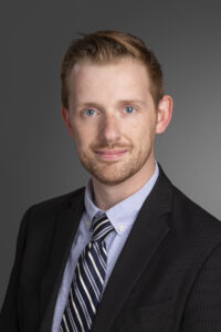 Professional headshot of Sean Watson with short light red hair and a trimmed beard, wearing a dark pinstripe suit, light blue collared shirt, and a navy striped tie. Sean is smiling slightly and posed against a neutral gray background