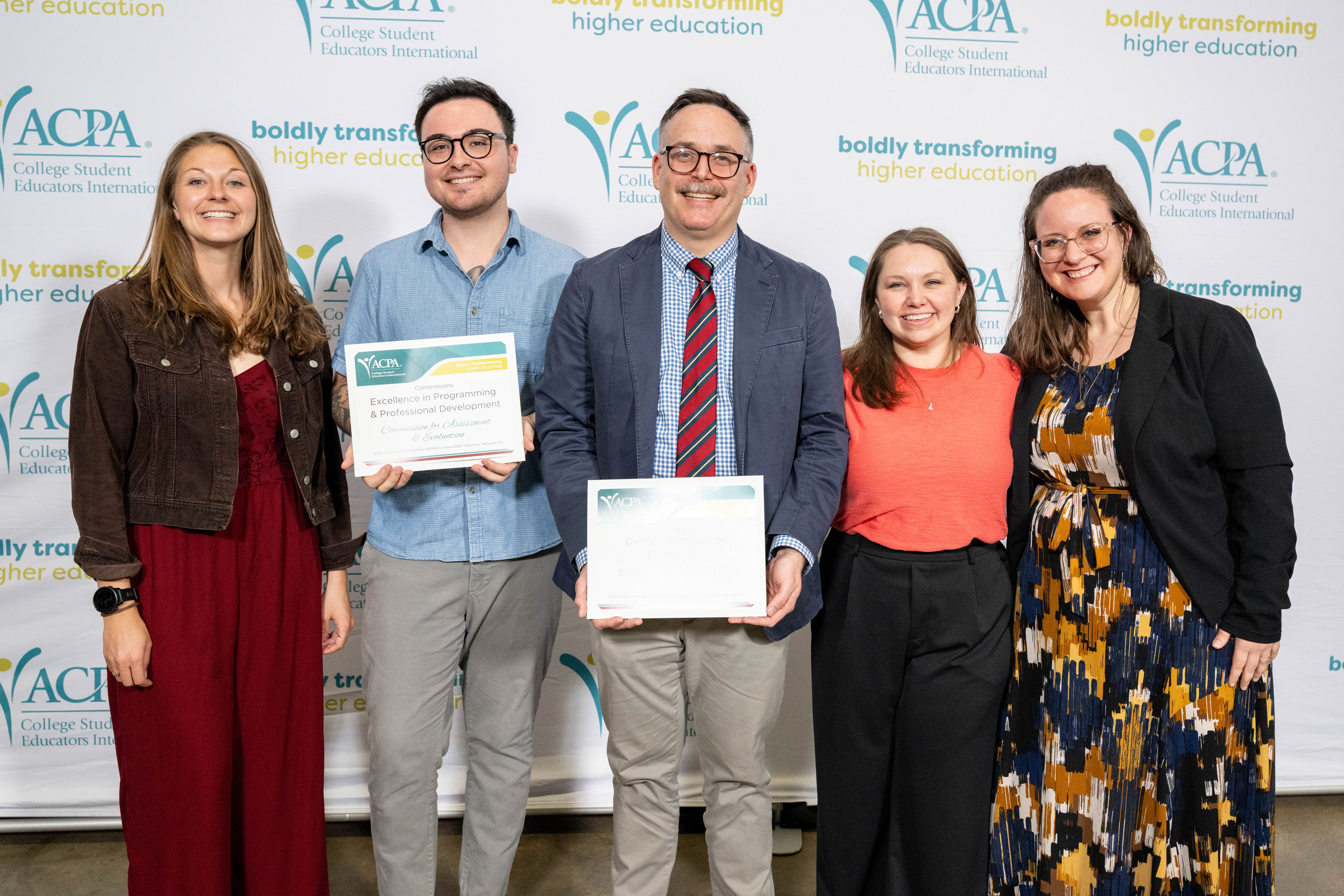  group of four people is posing in front of a backdrop that reads "ACPA College Student Educators International" with the slogan "boldly transforming higher education." Three of them are holding certificates. The woman on the left, wearing a white blazer, holds a certificate recognizing her as an "Annual Copolis Emerging Professional." The man next to her, wearing glasses and a suit, holds a certificate for "Distinguished Commission Directorate Member." The third person, wearing a dark suit and tie, holds a certificate for "Overall Distinguished Commission." The fourth individual, on the far right, is dressed in a sweater and lanyard. They are all smiling and looking at the camera.