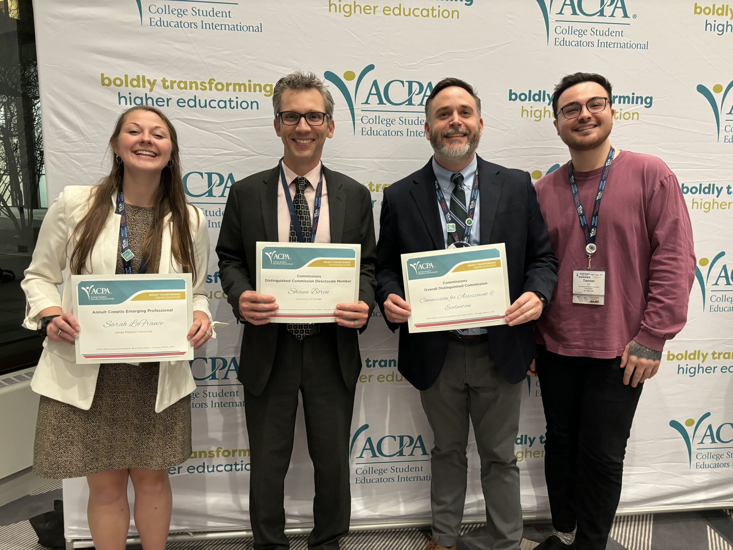  group of four people is posing in front of a backdrop that reads "ACPA College Student Educators International" with the slogan "boldly transforming higher education." Three of them are holding certificates. The woman on the left, wearing a white blazer, holds a certificate recognizing her as an "Annual Copolis Emerging Professional." The man next to her, wearing glasses and a suit, holds a certificate for "Distinguished Commission Directorate Member." The third person, wearing a dark suit and tie, holds a certificate for "Overall Distinguished Commission." The fourth individual, on the far right, is dressed in a sweater and lanyard. They are all smiling and looking at the camera.