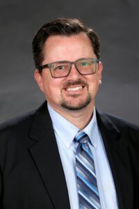 A headshot of George Still. A White man with glasses, wearing a suit and tie.