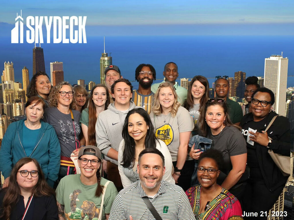 A group of 22 people stand in front of a view of the Chicago skyline. Everyone is smiling, and a few people are wearing shirts with their university's information. A logo in the top left corner identifies the location as Skydeck, located in downtown Chicago. The bottom right corner has a date of June 21, 2023.