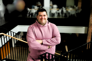 Photograph of a person wearing a pink hoodie standing with arms crossed on a staircase inside a modern building. Background shows blurred tables and chairs, indicating a casual or public indoor setting.