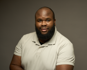 Portrait photo of a person with a shaved head and full beard wearing a light-colored polo shirt, seated against a dark gray background and looking toward the camera with a slight smile. The image is a professional head-and-shoulders style photograph with soft lighting.