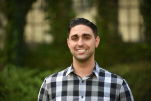 Photograph of a person wearing a black and white checkered shirt standing outdoors with blurred greenery and a building in the background. The person's face is obscured by a blur.