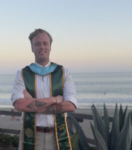 Photograph of a person wearing a graduation stole standing with arms crossed near a beach at sunset. Stole features green and gold colors with "Cal Poly 2023" embroidered, and person has visible tattoos on forearms.