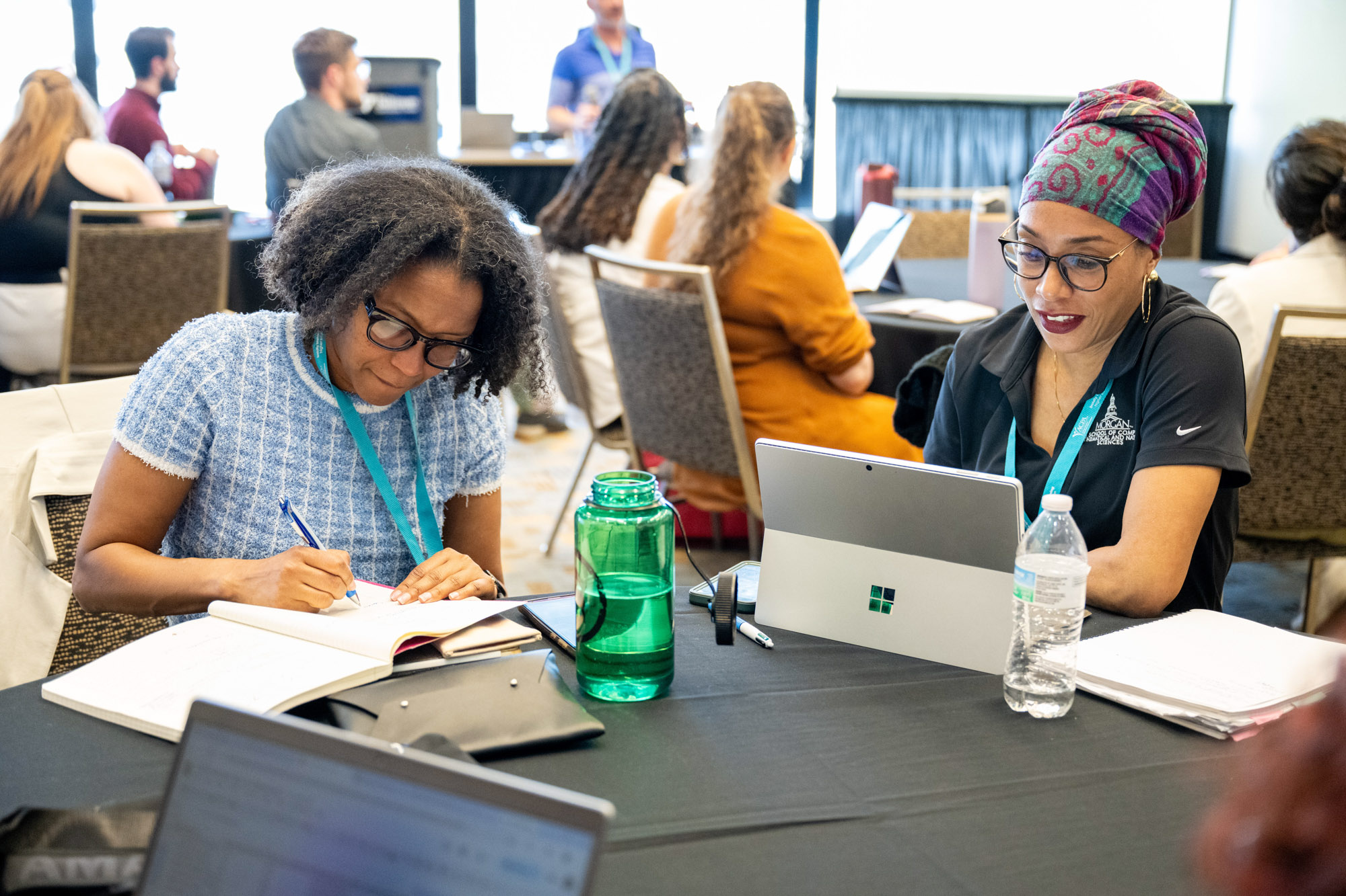 two individuals sitting at a table looking at computers and notepads taking notes in a room during a presentation